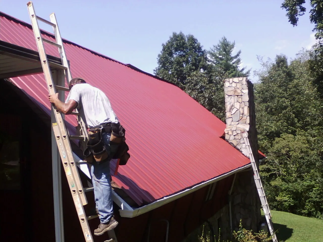 Roofer on a ladder beside a red metal roof and stone chimney — Soffit & Fascia Repair by Roofing Brothers in Eola